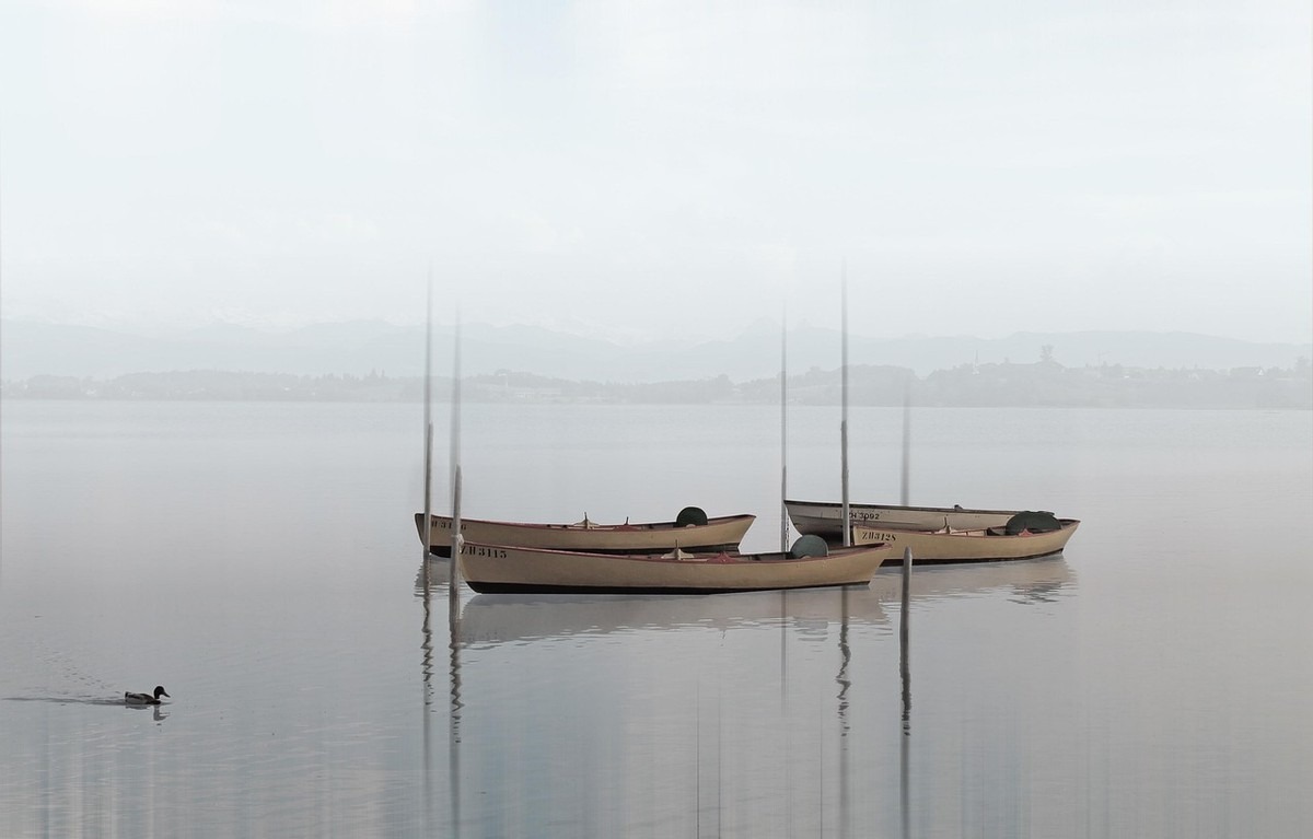 Trois petits bateaux de pêche amarrés dans un lac brumeux par temps calme, avec des montagnes floues en arrière-plan