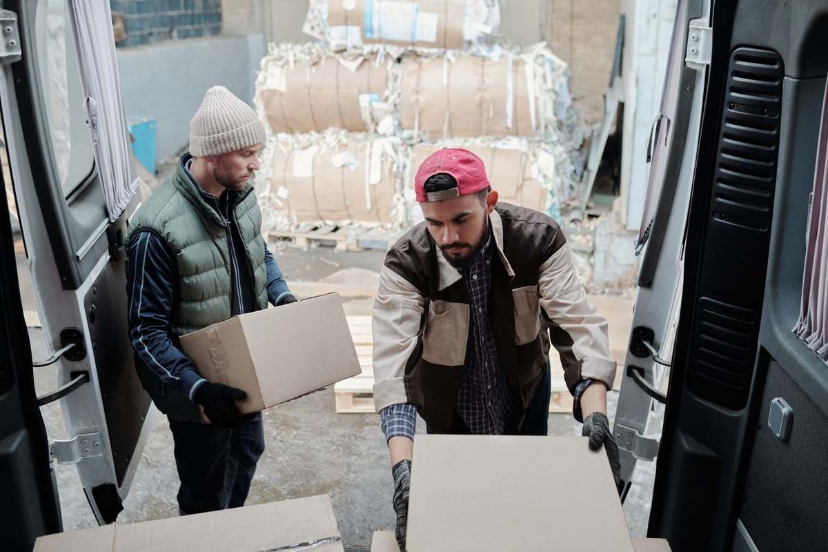 Homme en rouge décharge un carton, autre homme observe, camion ouvert, entrepôt en arrière-plan